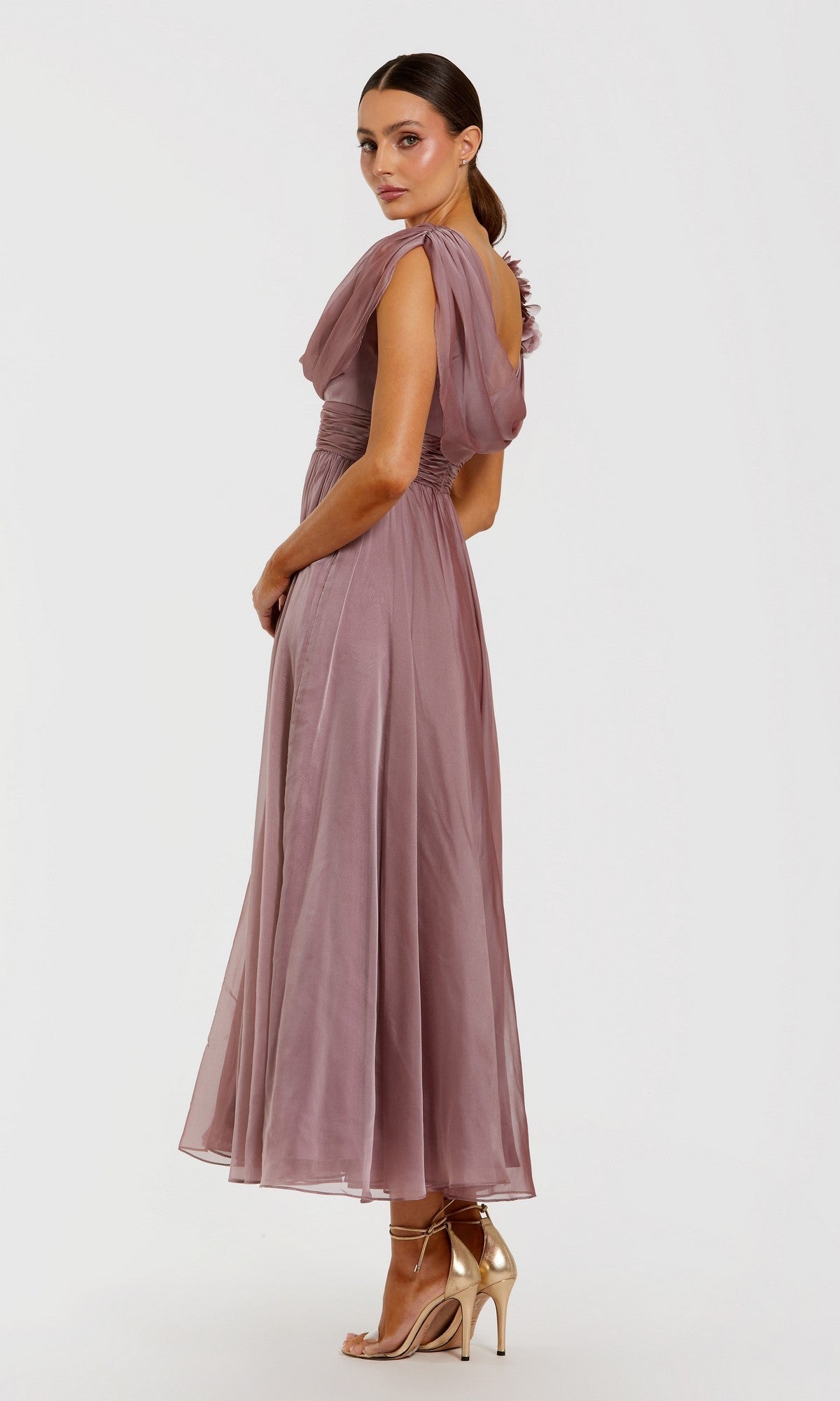 A woman wearing the Mac Duggal 68967 Tea-Length Party Dress—crafted from mauve, flowy chiffon with a one-shoulder neckline and ruched waist detail—paired with gold heeled sandals, stands against a plain white background.